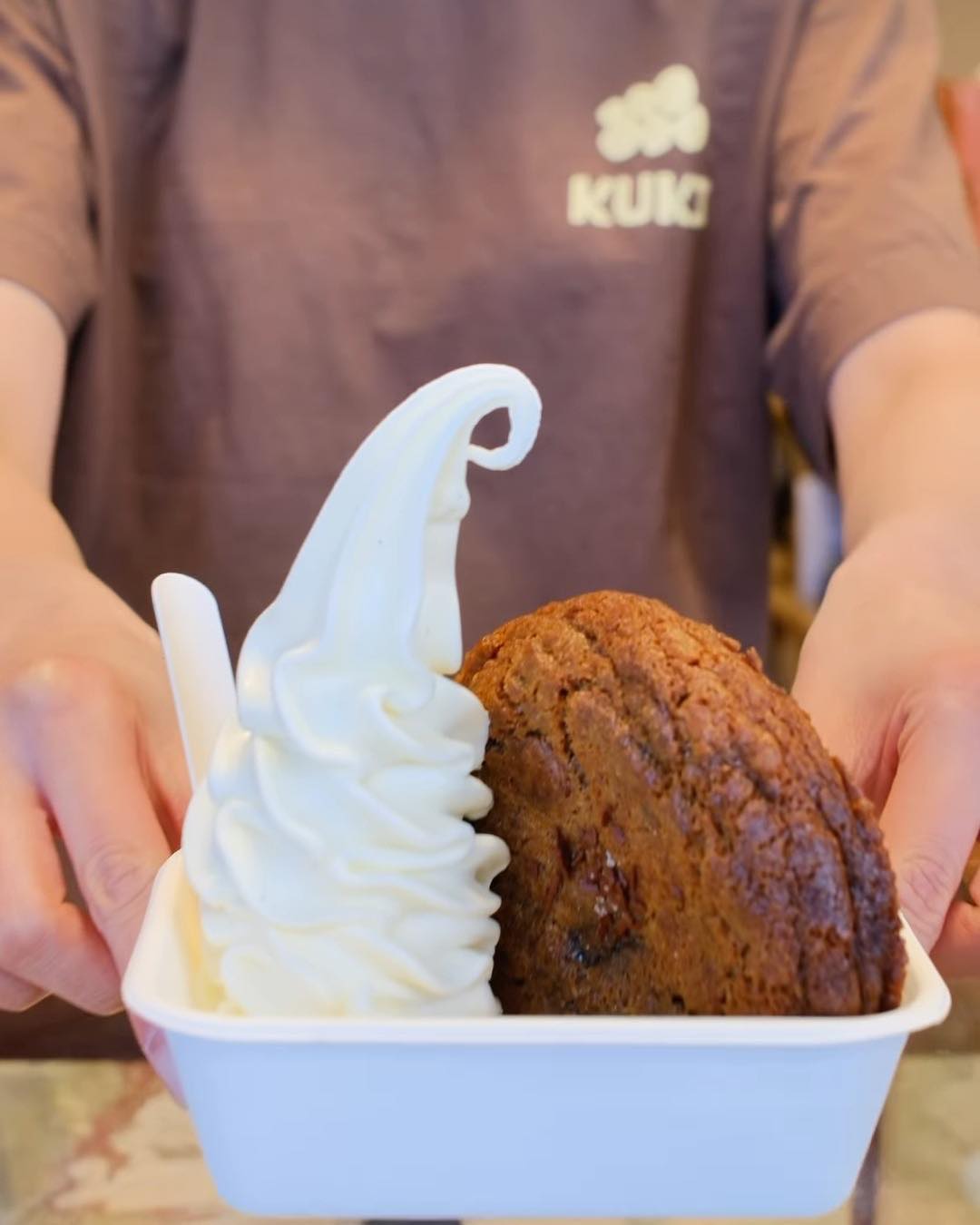 A KUKI staff member in a brown KUKI tee holds out a soft-serve twirl and a cookie in a white tray.