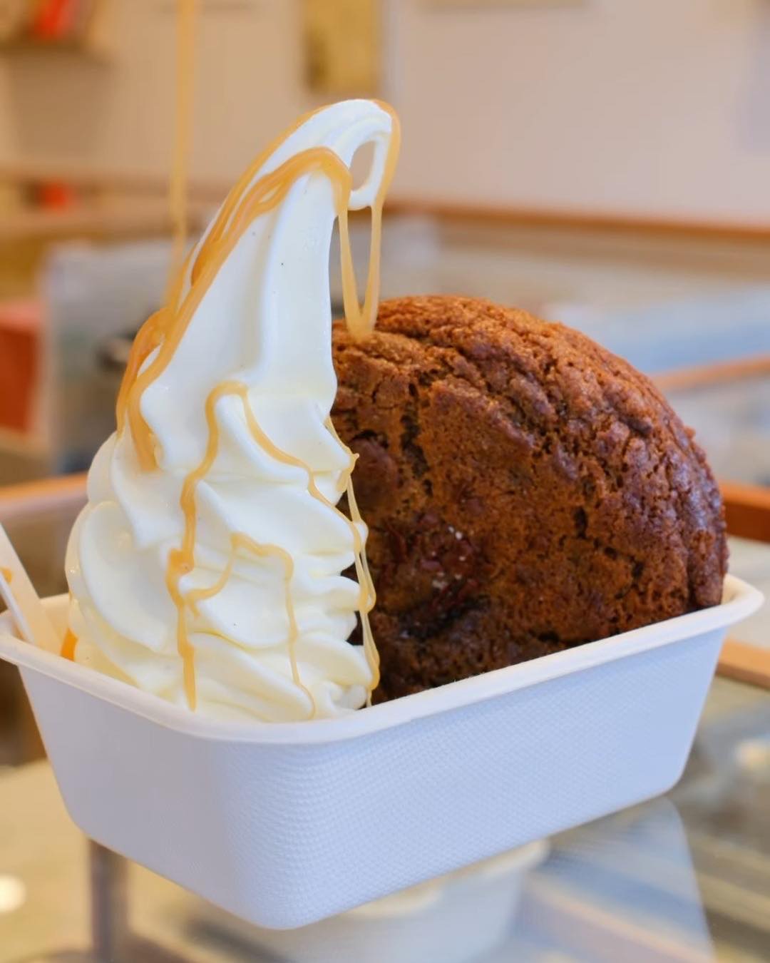 A KUKI soft-serve twirl with a caramel drizzle beside a whole cookie in a paper tray.