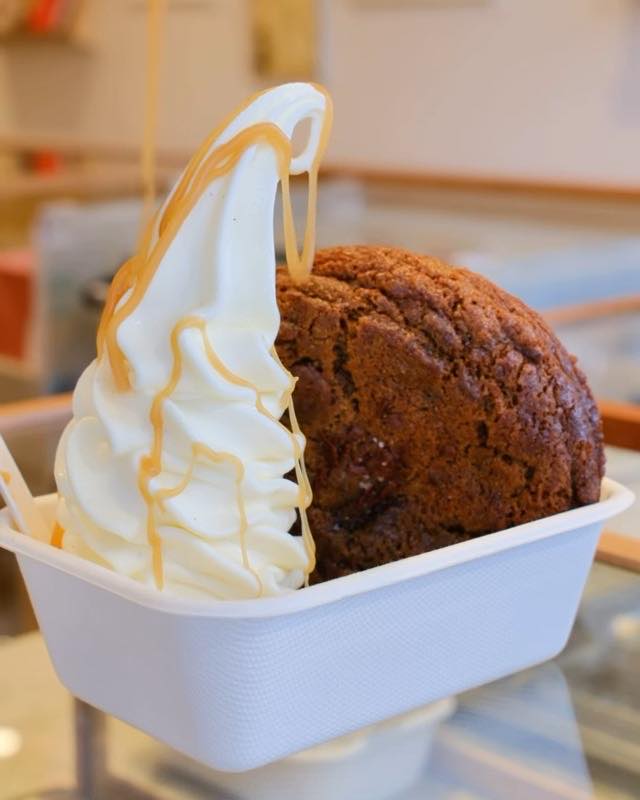 A KUKI soft-serve twirl with a caramel drizzle beside a whole cookie in a paper tray.